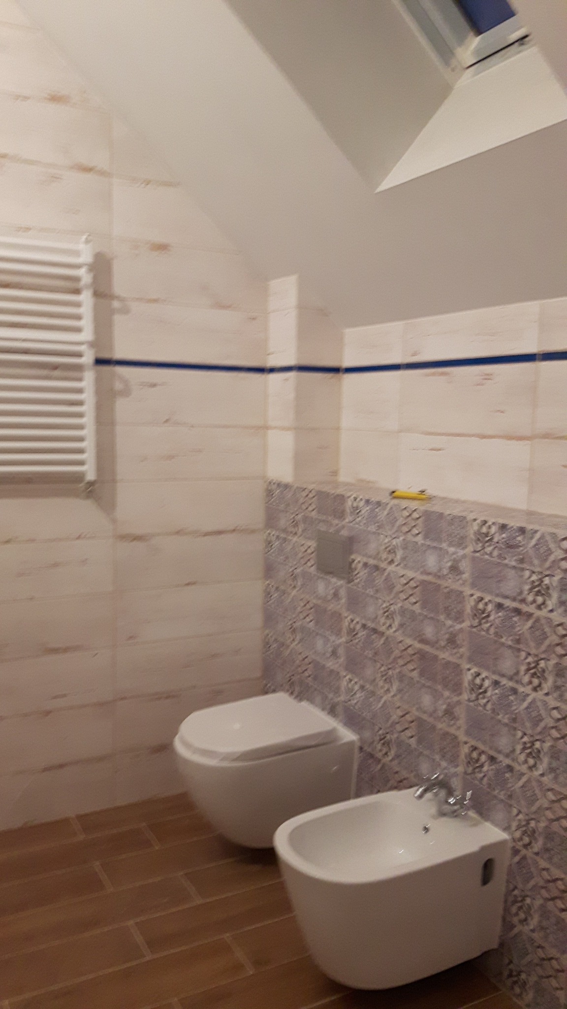 Wykończona łazienka na poddaszu z toaletą podwieszaną, bidetem i grzejnikiem drabinkowym, wyłożona płytkami w dwóch wzorach z niebieskim akcentem.