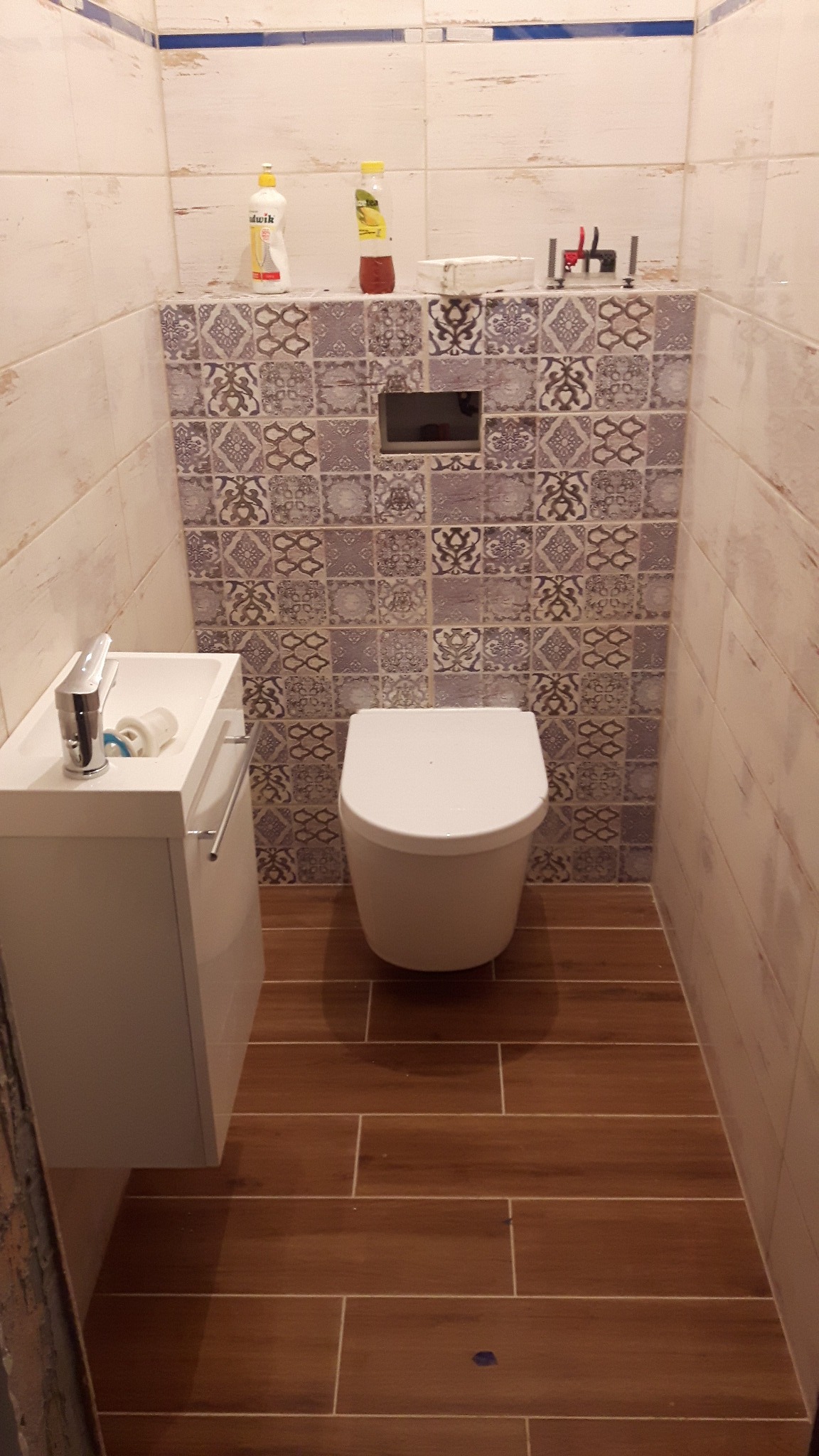 Nowoczesna toaleta w trakcie wykańczania: biała miska WC, umywalka z szafką, dekoracyjne płytki ścienne z wzorem i imitujące drewno płytki podłogowe, widoczne akcesoria montażowe na ścianie.