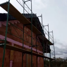 Dom w trakcie budowy z widocznymi rusztowaniami, ociepleniem ścian i fragmentem dachu pokrytego dachówką, na tle pochmurnego nieba.