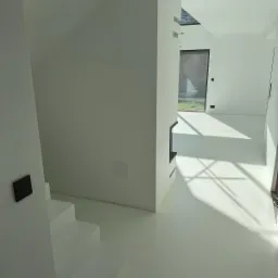 Minimalistyczne wnętrze: białe schody prowadzą na gładką, jednolitą posadzkę w jasnym odcieniu, z widocznymi promieniami słońca wpadającymi przez okno.