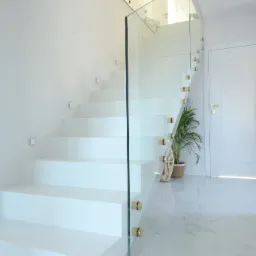Nowoczesne, białe schody betonowe z szklaną balustradą mocowaną na złotych elementach, oświetlone małymi kwadratowymi lampkami w ścianie, z rośliną doniczkową u podnóża i marmurową podłogą.
