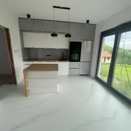 Biała kuchnia z wyspą i marmurkową posadzką żywiczną, widok na ogród przez duże okno.