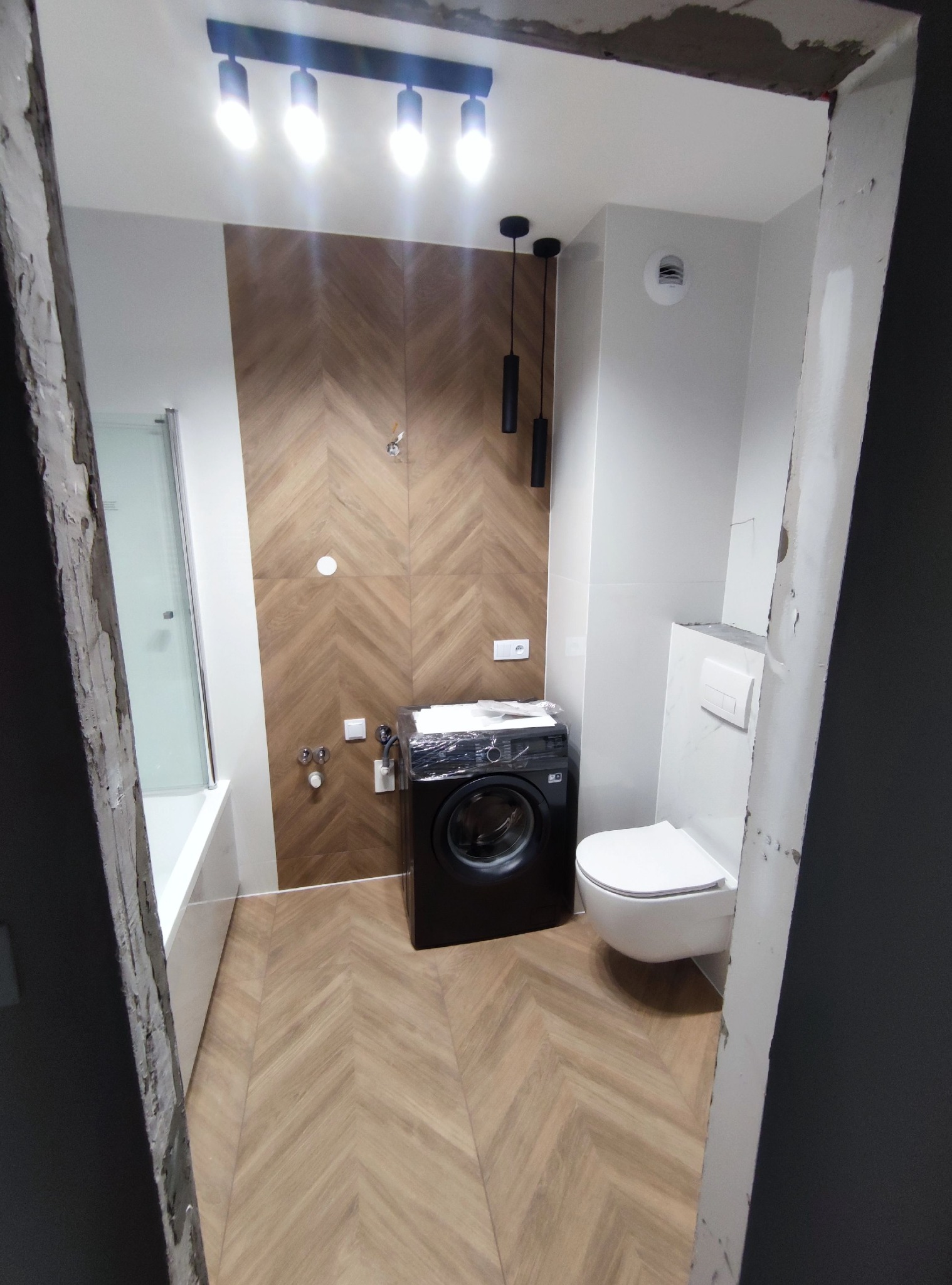 Nowoczesna łazienka w trakcie wykańczania, z drewnopodobnymi płytkami ułożonymi w jodełkę na podłodze i ścianie, białą armaturą i czarną pralką w folii ochronnej, widok zza surowego otworu drzwiowego.