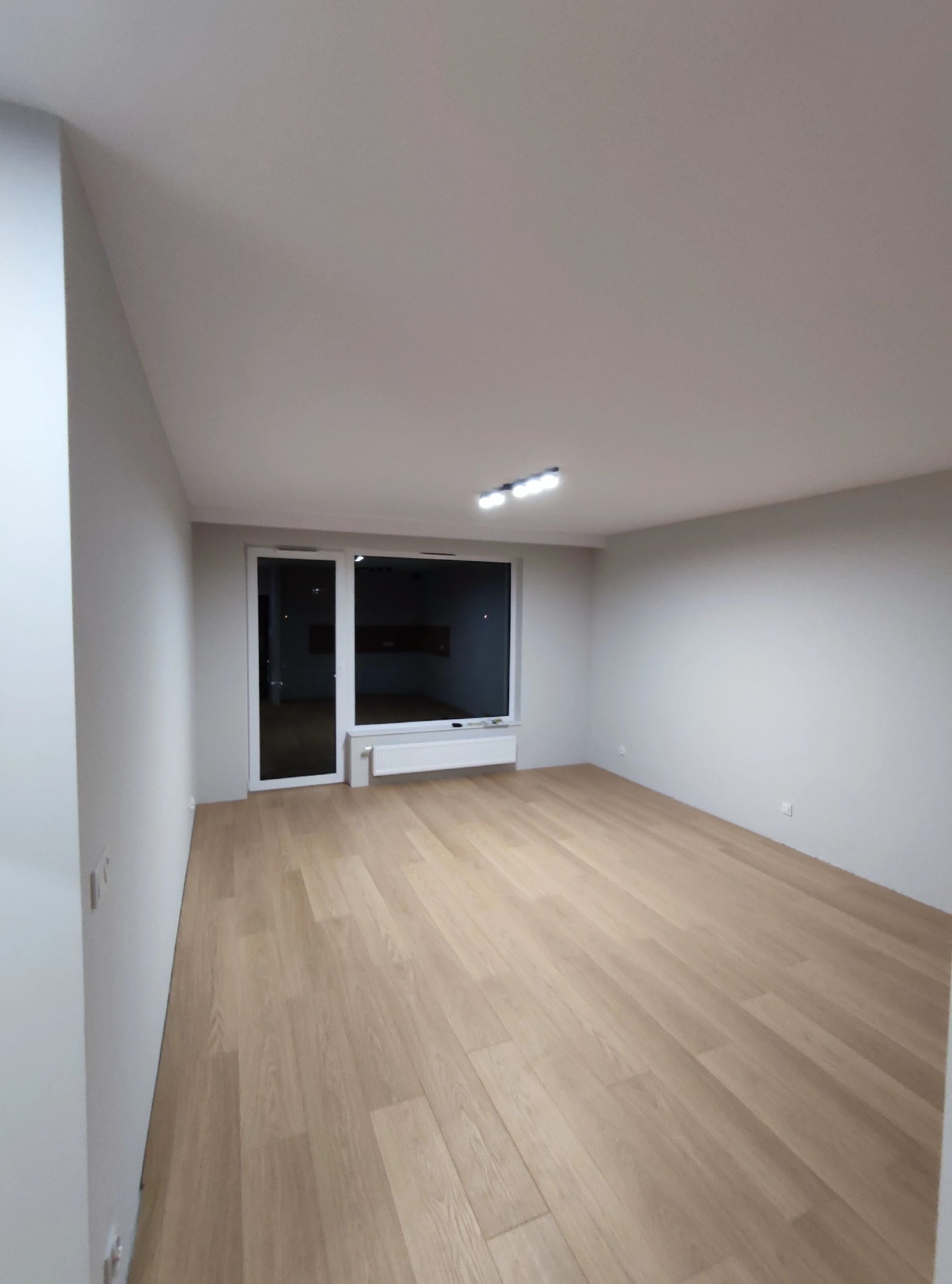 Pusty, jasny pokój po remoncie w Grójcu, z drewnianą podłogą i świeżo pomalowanymi na szaro ścianami. Widoczne białe okno z grzejnikiem i nowoczesne oświetlenie sufitowe.