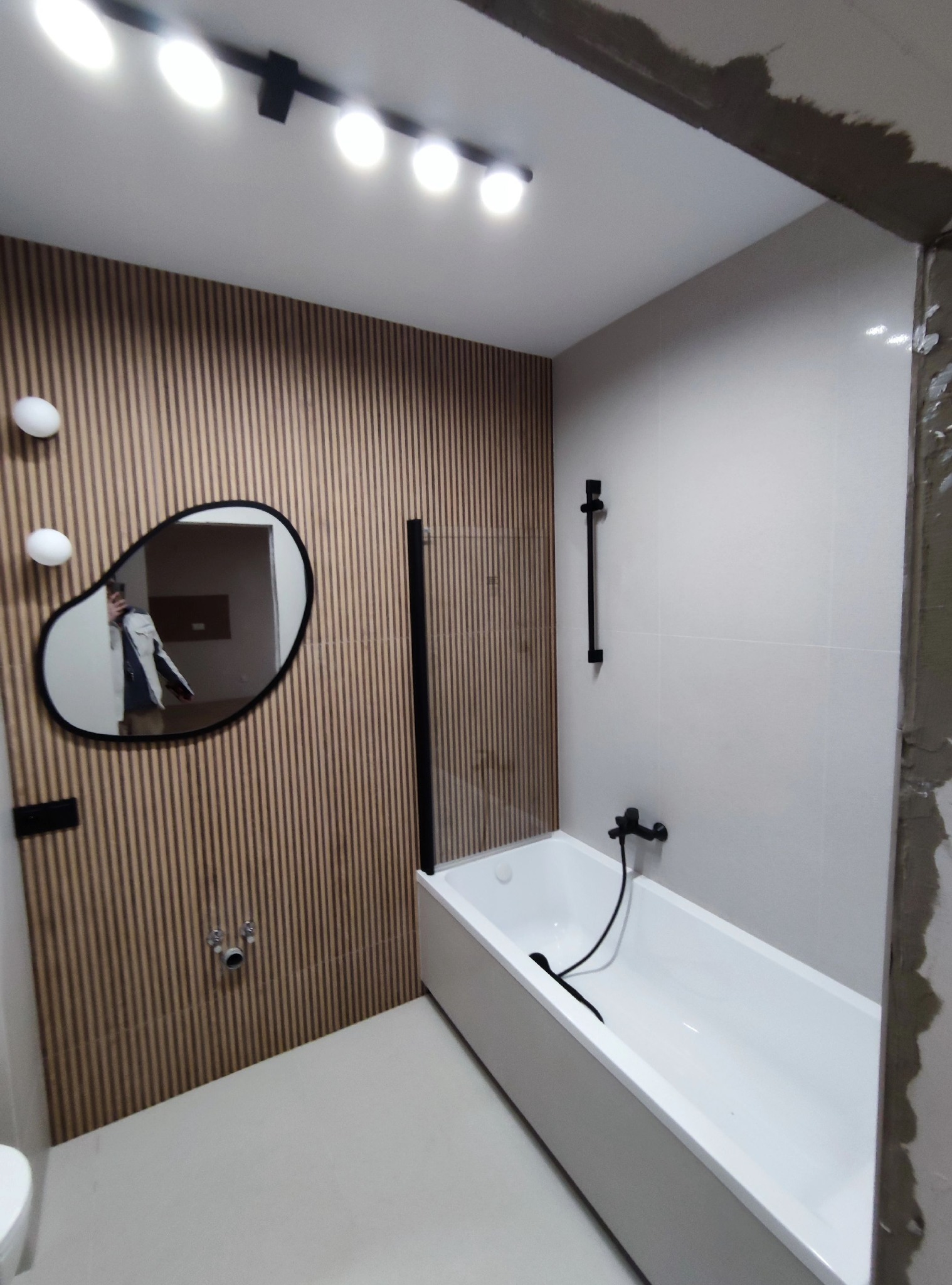 Nowoczesna łazienka z wanną, drewnianą ścianą z lamelami i asymetrycznym lustrem, w którym odbija się osoba w kurtce.