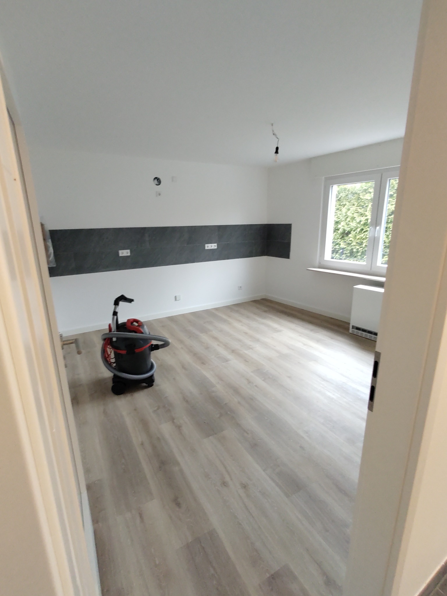 Pusty pokój po remoncie z nową podłogą, pomalowanymi ścianami z szarym pasem, odkurzacz przemysłowy w rogu, widok zza otwartych drzwi.