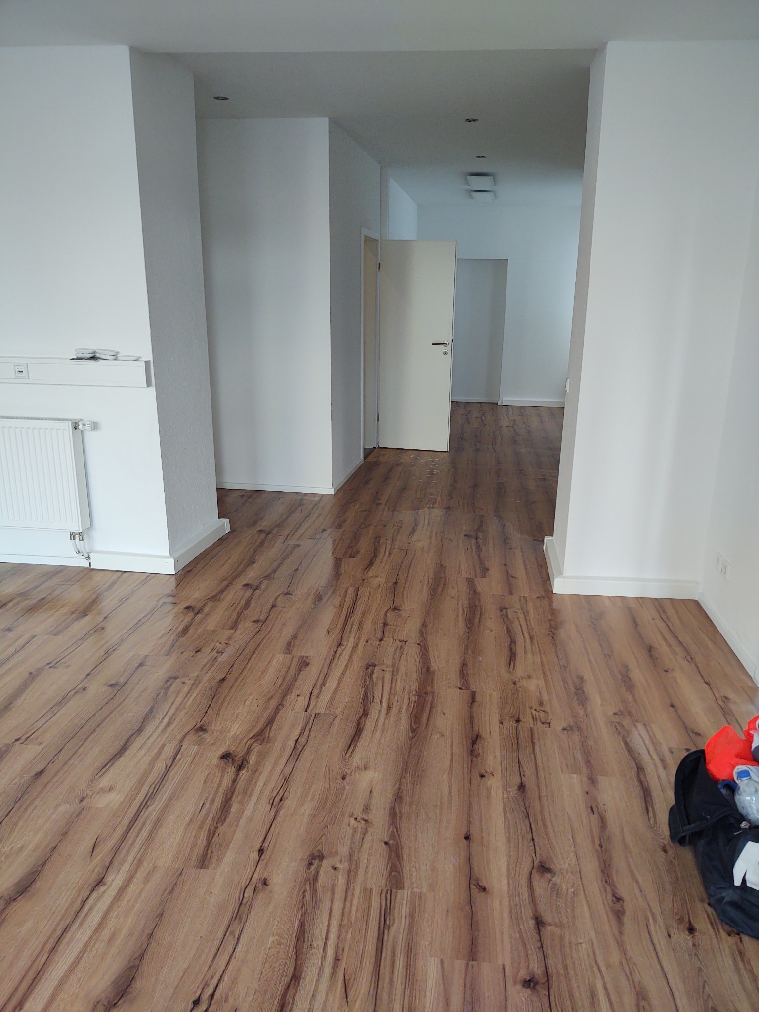 Puste, świeżo wykończone wnętrze z panelami podłogowymi o wzorze drewna, białe ściany i drzwi, widoczny grzejnik i torba z narzędziami w rogu.