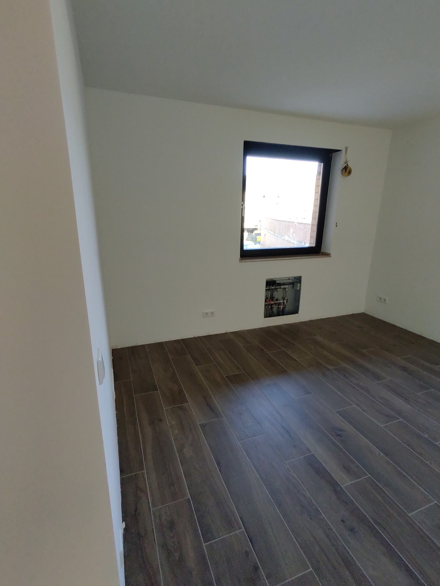 Pusty pokój z brązowymi płytkami podłogowymi imitującymi drewno, białymi ścianami i oknem z czarną ramą; widoczna instalacja w ścianie pod oknem.