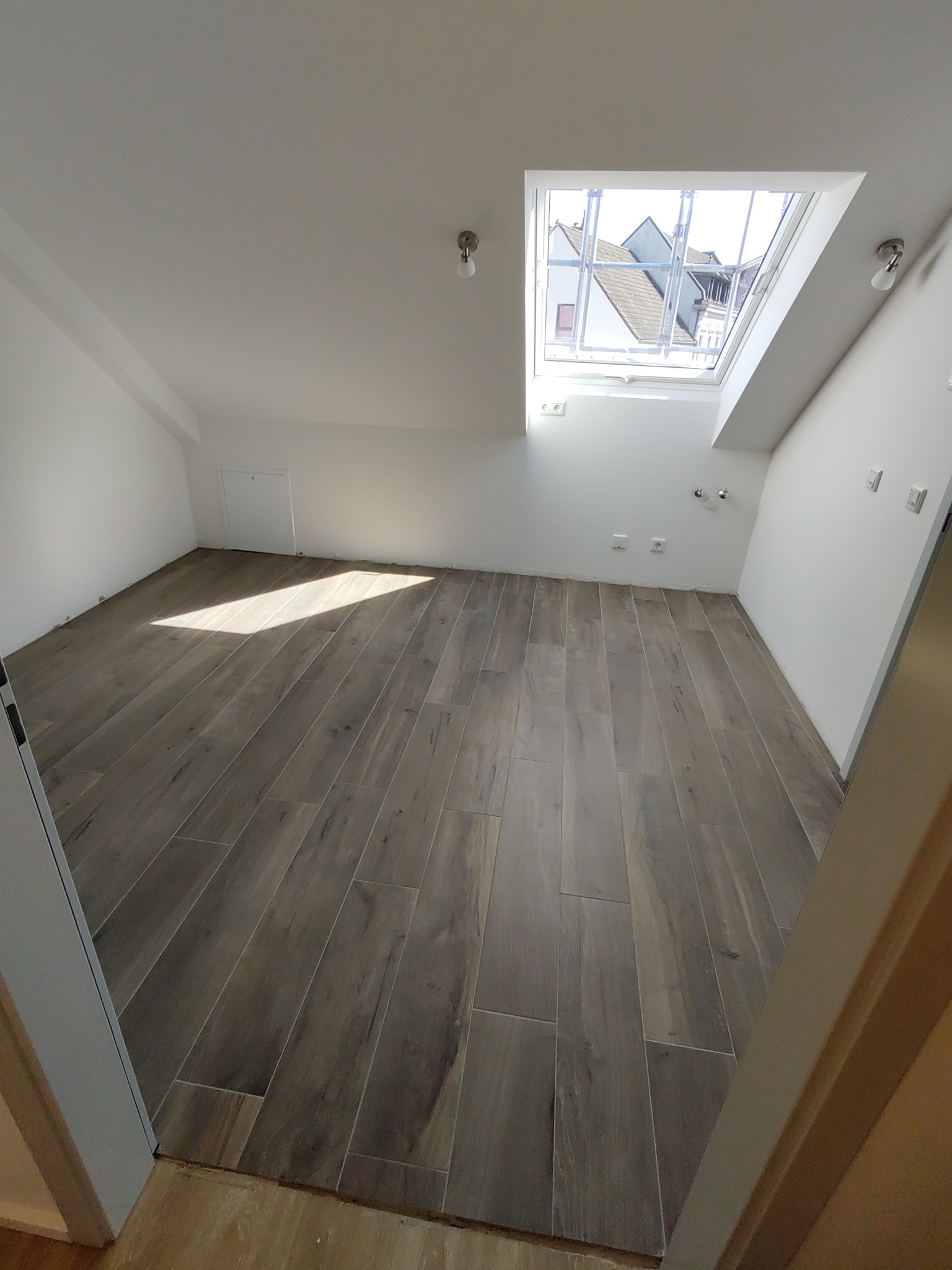 Puste poddasze z drewnopodobną podłogą z paneli, oświetlone światłem wpadającym przez okno dachowe; świeżo pomalowane białe ściany i sufit.