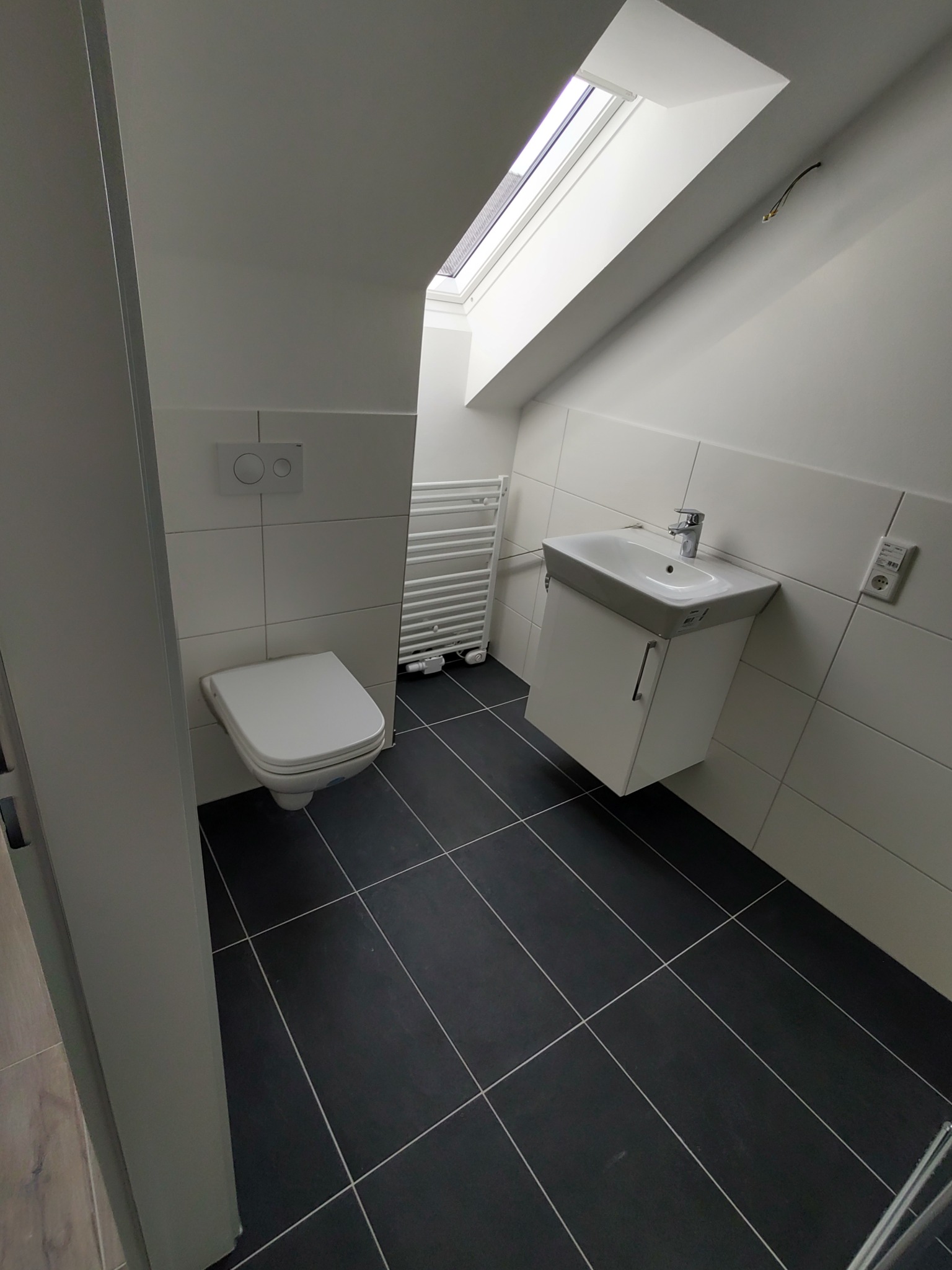 Nowoczesna, minimalistyczna łazienka na poddaszu z czarną podłogą, białymi ścianami, toaletą, grzejnikiem drabinkowym i umywalką z szafką, oświetlona światłem z okna dachowego.