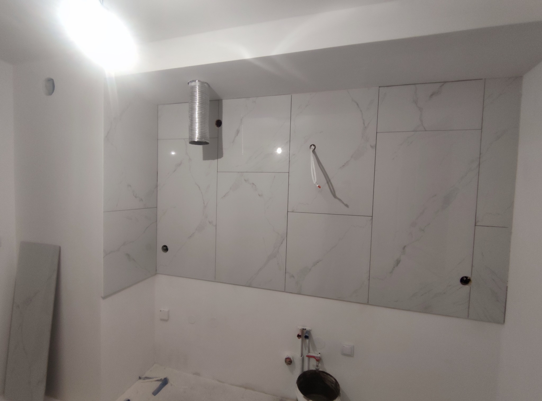 Świeżo wykafelkowana ściana w łazience z marmurowym wzorem, widoczny fragment instalacji wentylacyjnej i przygotowane przyłącza wodne.