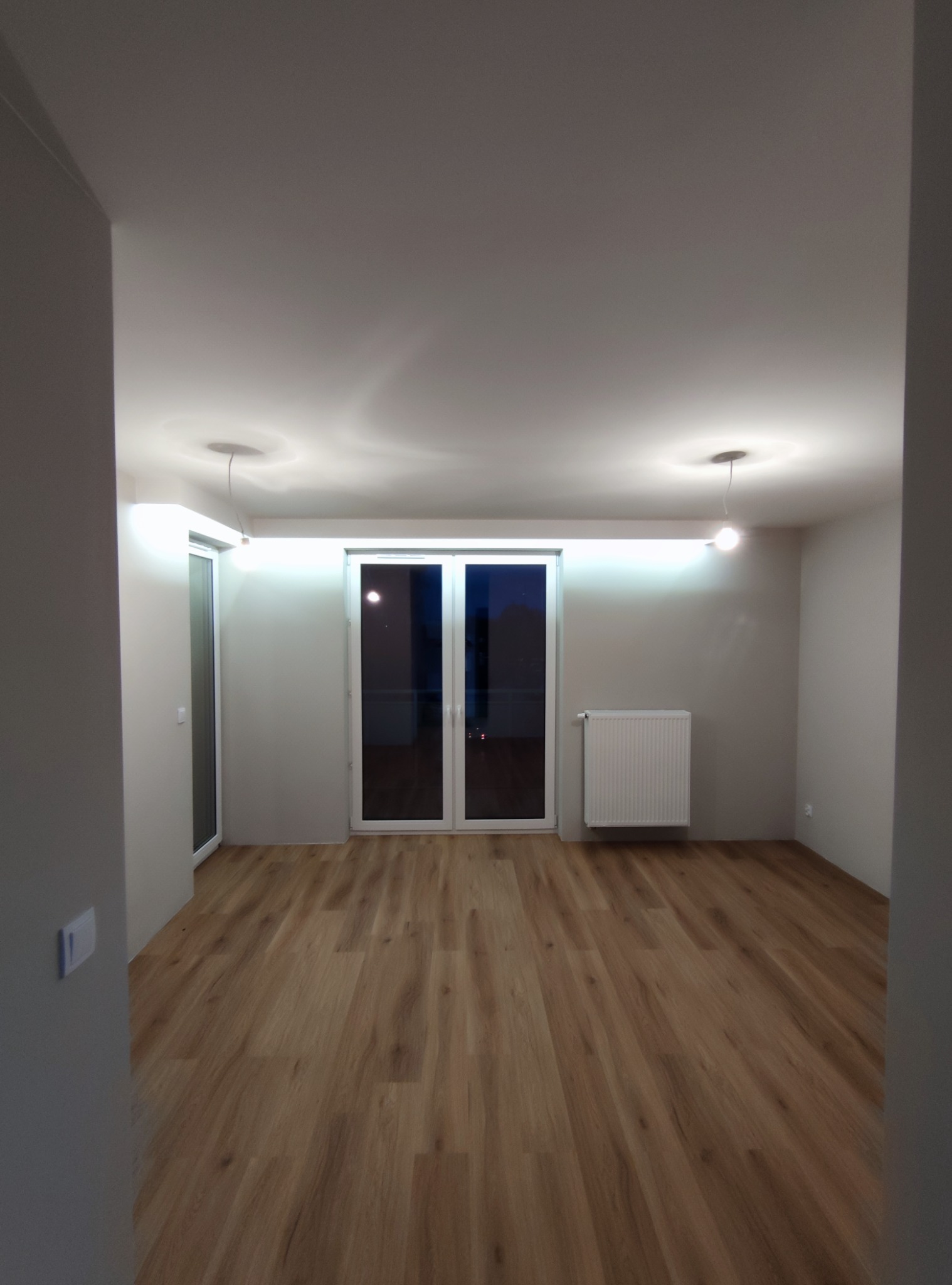 Puste, jasne wnętrze z panelami podłogowymi, białymi ścianami, grzejnikiem i oknami balkonowymi, oświetlone wiszącymi lampami i listwą LED nad oknem.