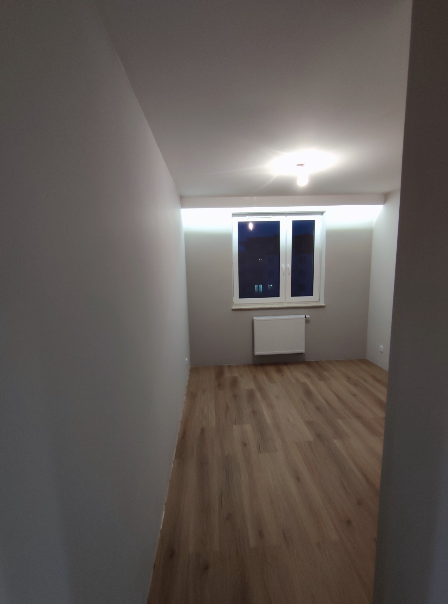 Pomalowane na szaro wnętrze pokoju z drewnianą podłogą, białym oknem i grzejnikiem, widoczne z perspektywy wejścia.