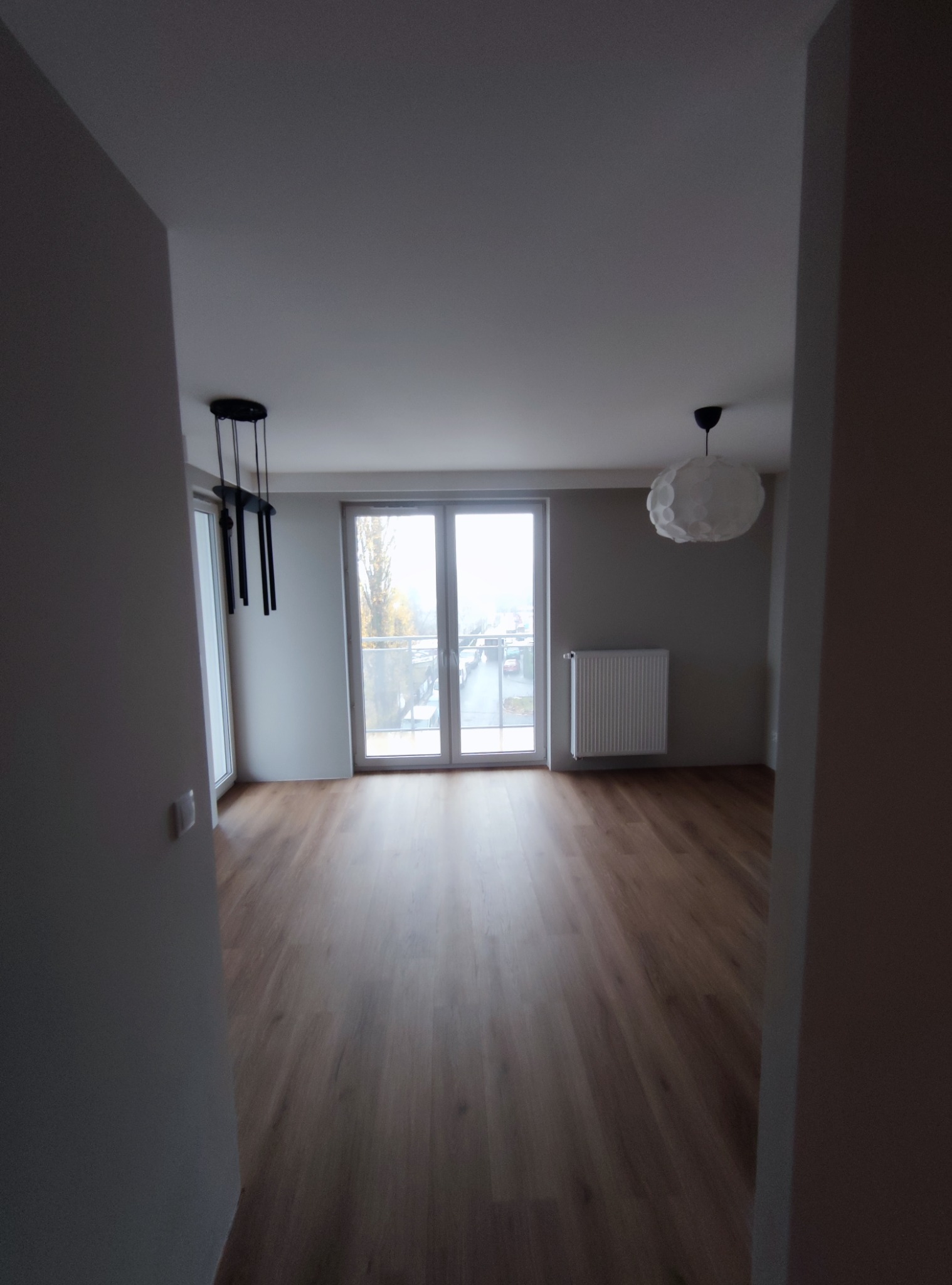 Pusty pokój z drewnianą podłogą, białymi ścianami i grzejnikiem pod oknem balkonowym, widok z korytarza, dwie różne lampy sufitowe.