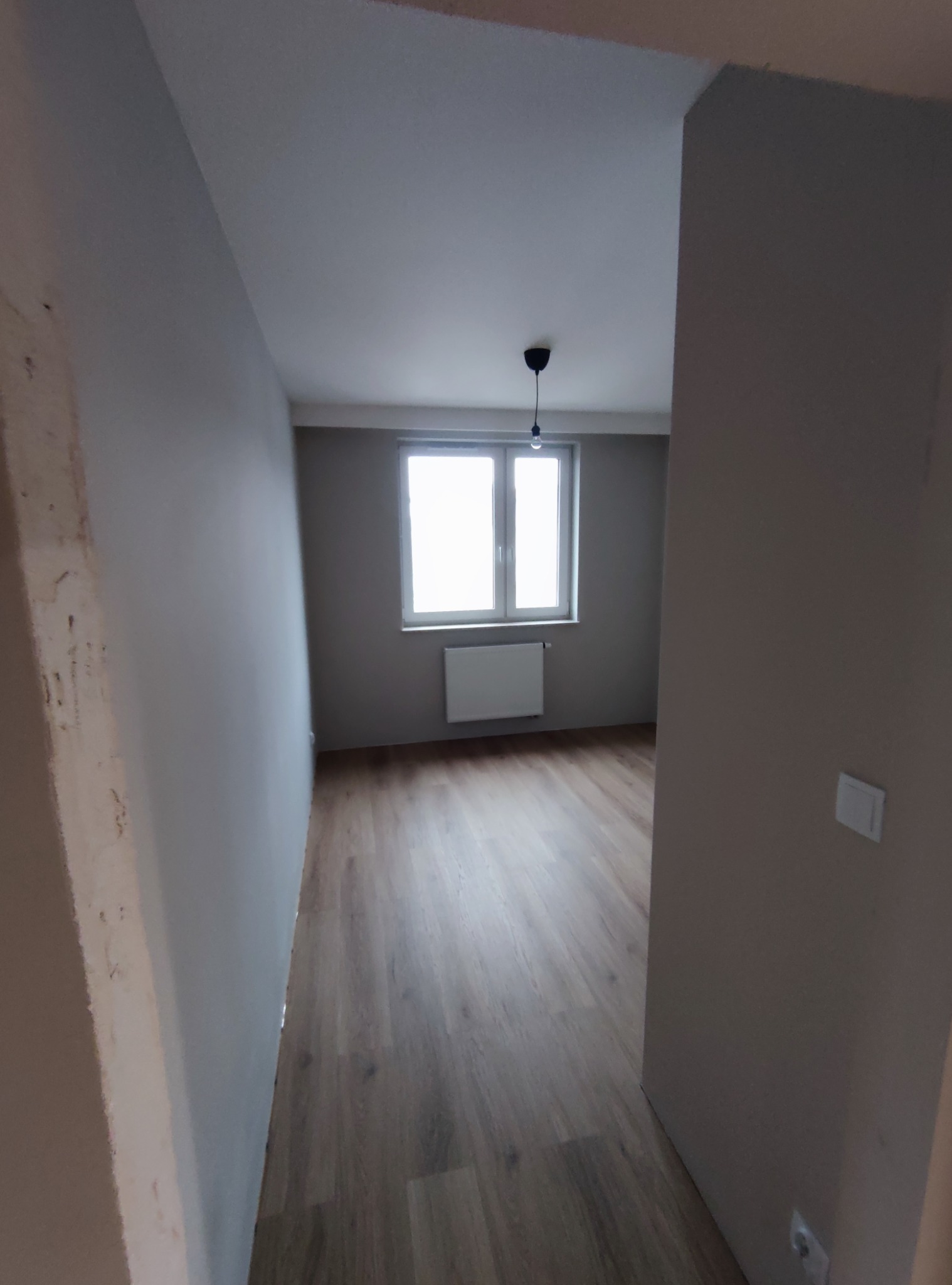 Pomalowane na szaro wnętrze pokoju z panelami podłogowymi, widok z korytarza, ściana po lewej stronie w trakcie wykańczania.