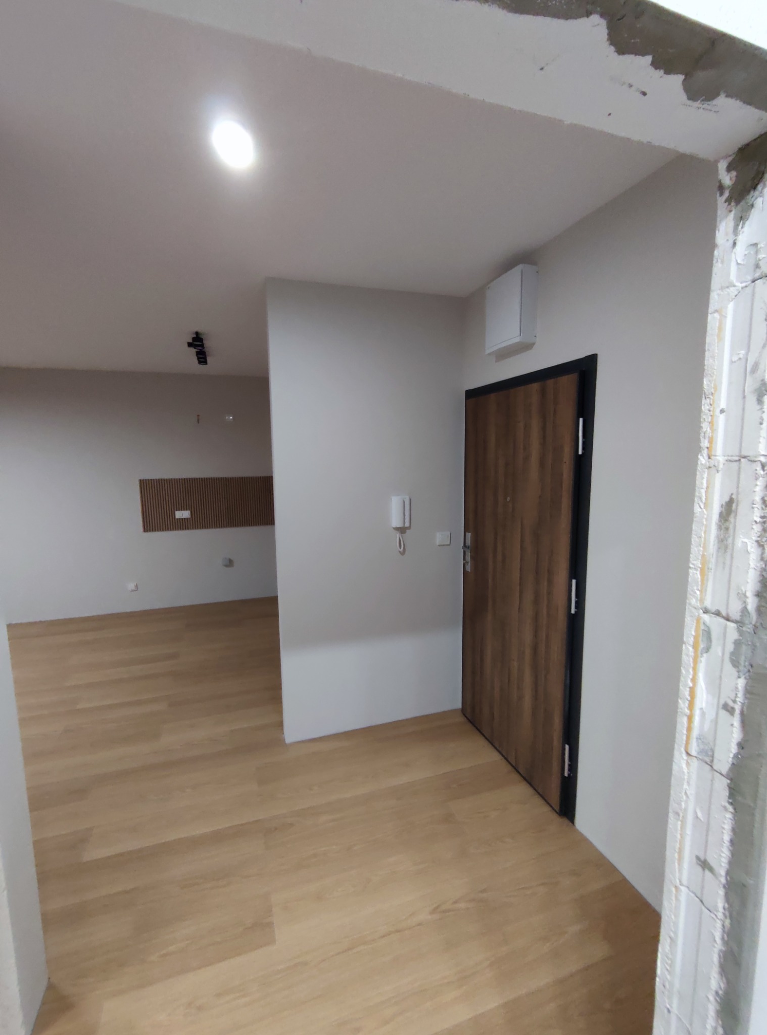 Wnętrze mieszkania w trakcie wykańczania: świeżo pomalowane ściany w odcieniach bieli, drewniana podłoga, drzwi wejściowe z czarną ościeżnicą i widoczne ślady prac wykończeniowych na futrynie.