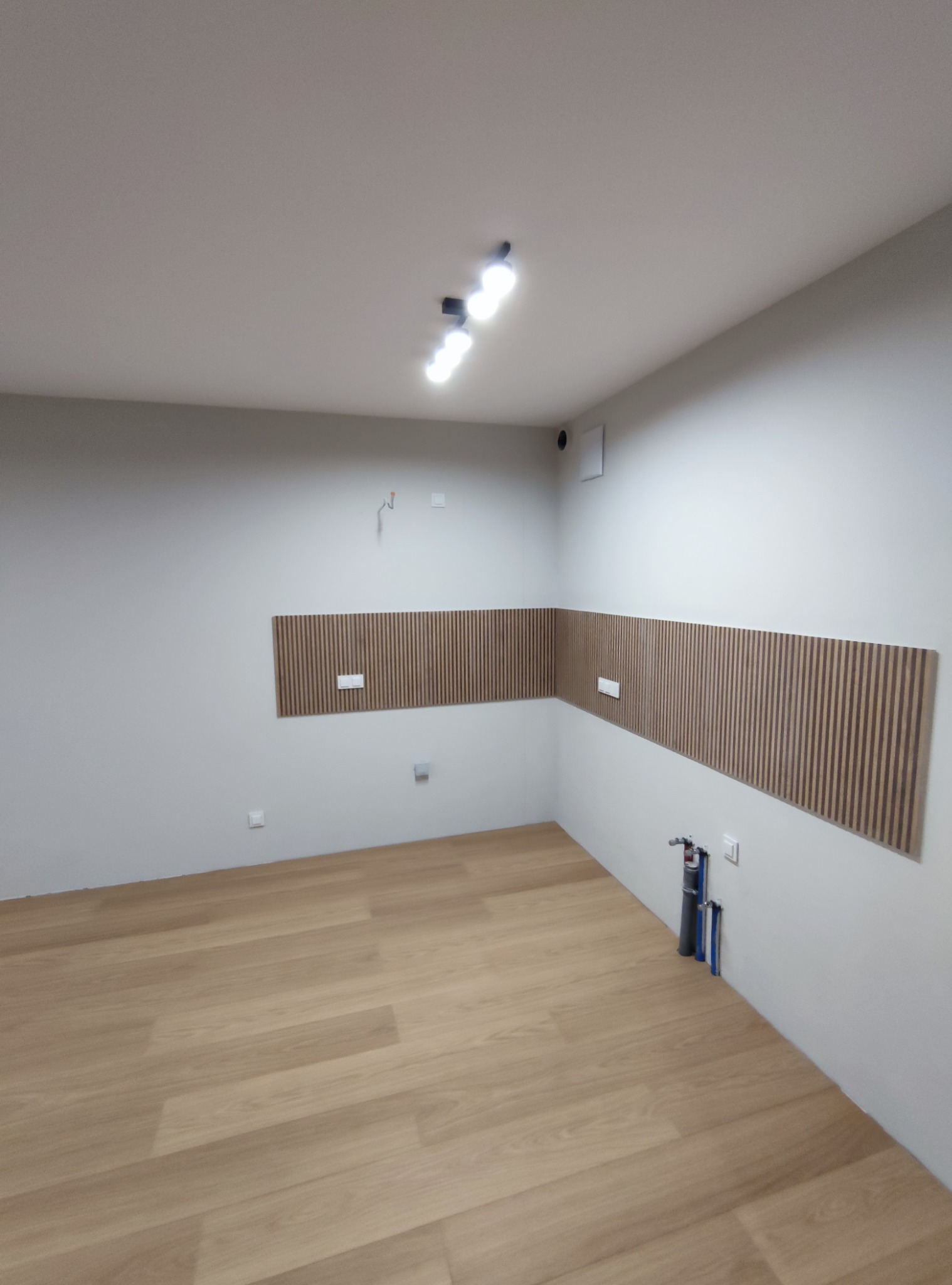Puste pomieszczenie po remoncie z drewnianą podłogą i panelami ściennymi z pionowych listewek, widoczne wystające rury instalacji wodnej.