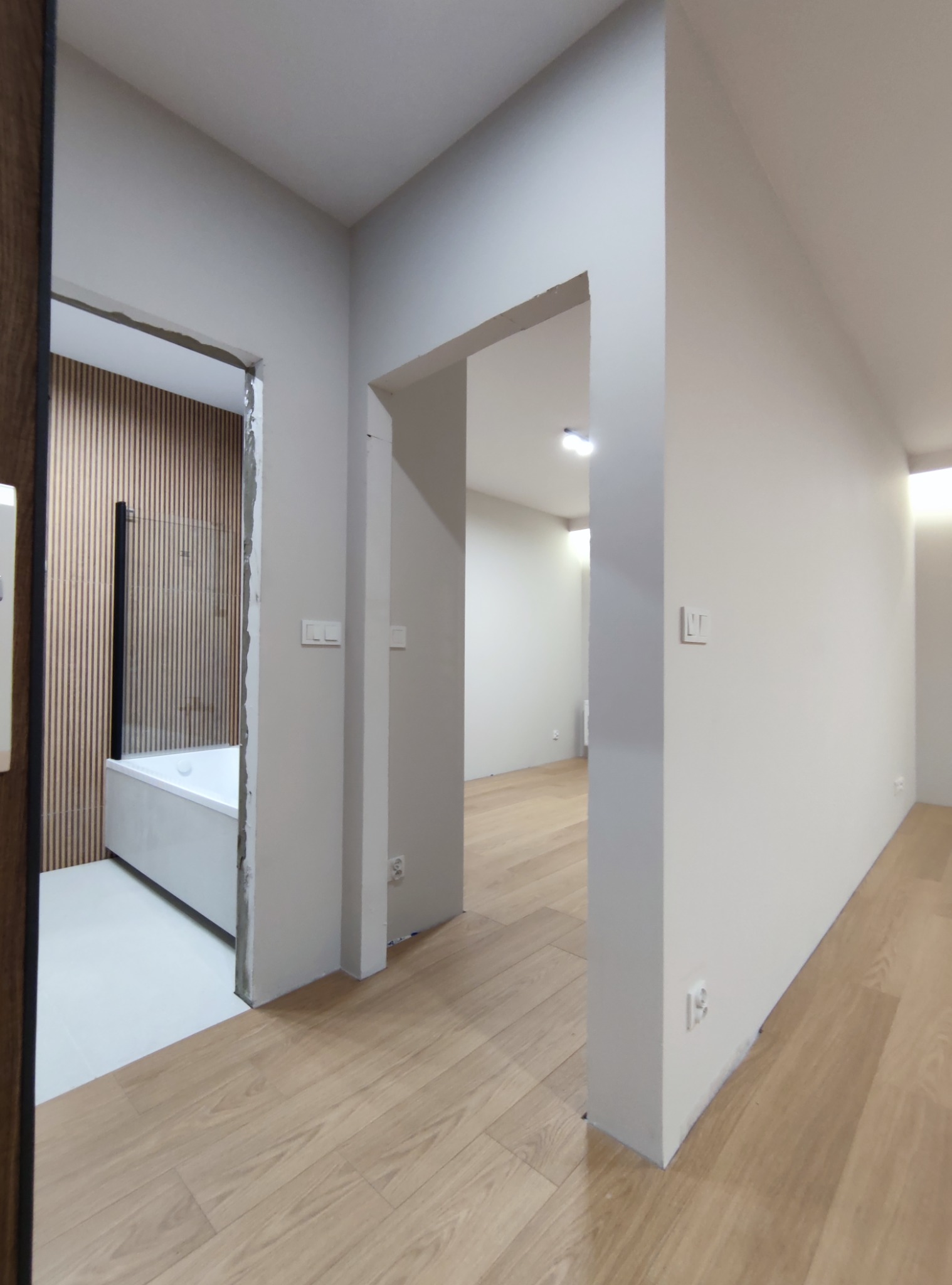 Wnętrze mieszkania w trakcie wykańczania: widok na łazienkę z drewnianymi panelami i wanną przez niedokończone przejście, jasne ściany i drewnopodobna podłoga w korytarzu.