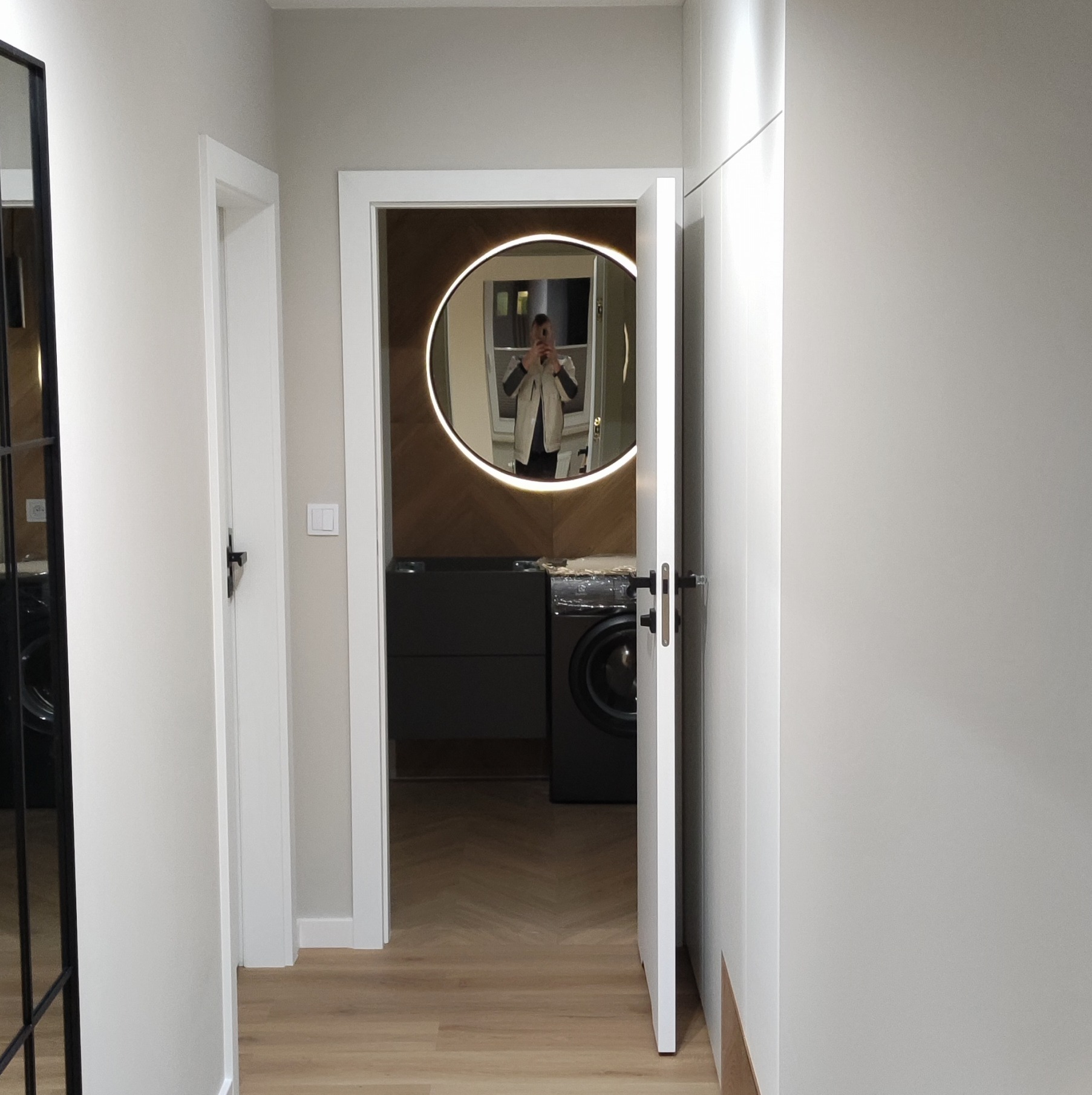 Nowoczesna łazienka z okrągłym lustrem podświetlanym LED, widoczna przez otwarte drzwi z korytarza, z pralką i szafką pod umywalkę w kolorze grafitowym oraz parkietem ułożonym w jodełkę.
