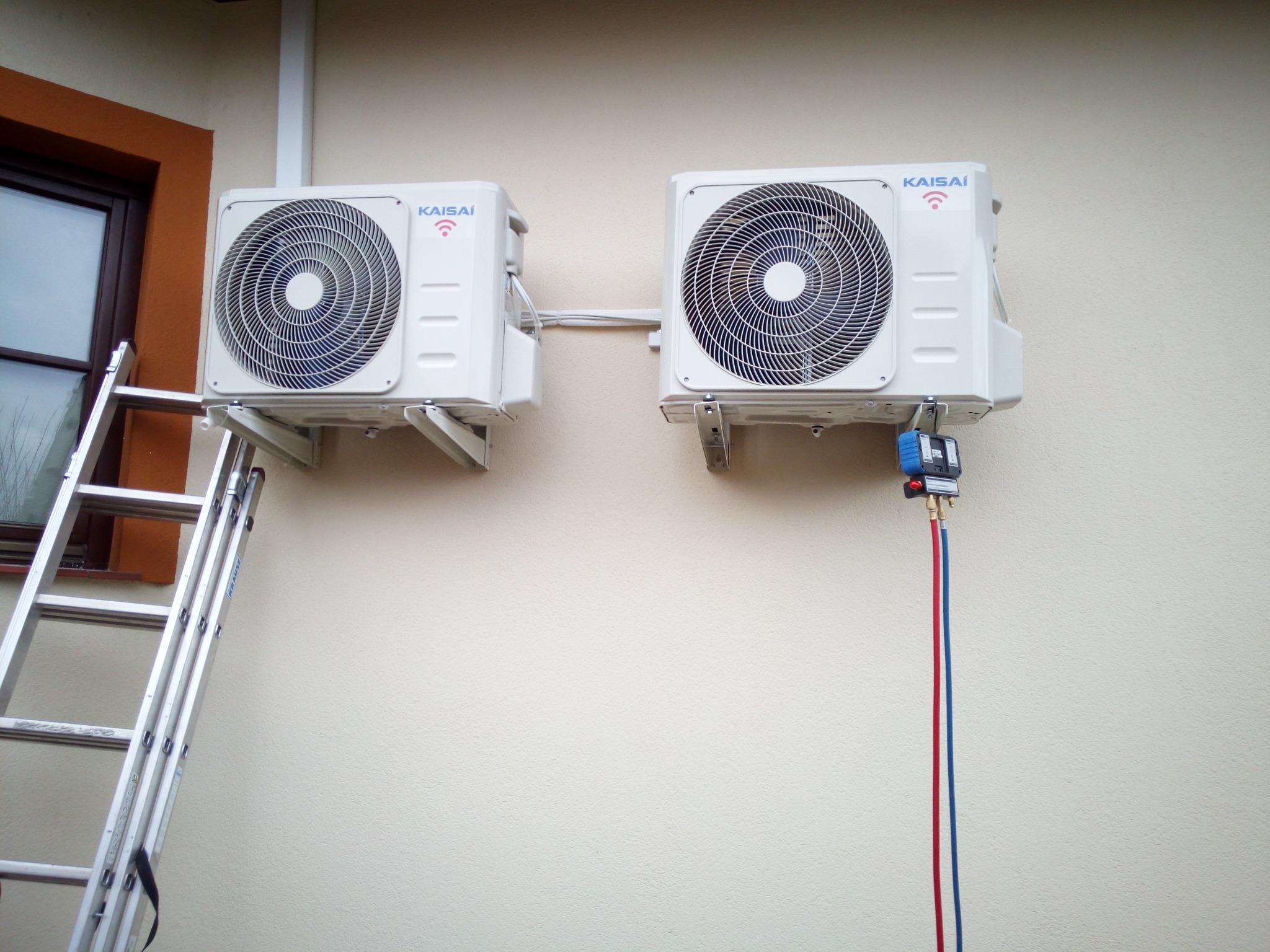 Dwa białe klimatyzatory marki Kaisai zamontowane na ścianie budynku, podłączone do narzędzia serwisowego z czerwonym i niebieskim przewodem, obok drabina aluminiowa