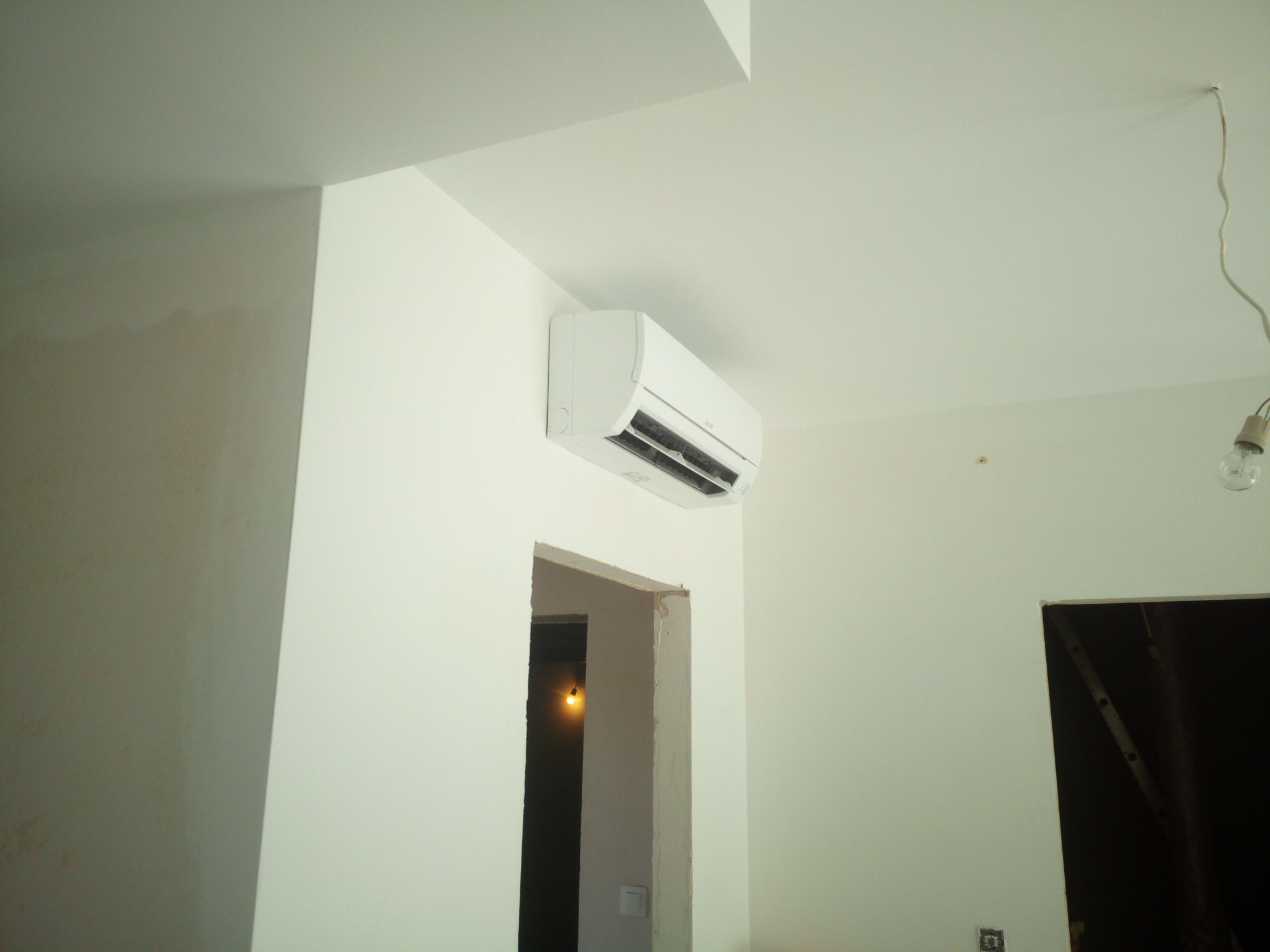 Biały klimatyzator ścienny zamontowany w jasnym pomieszczeniu, widoczny fragment sufitu, drzwi w stanie surowym oraz wisząca żarówka.