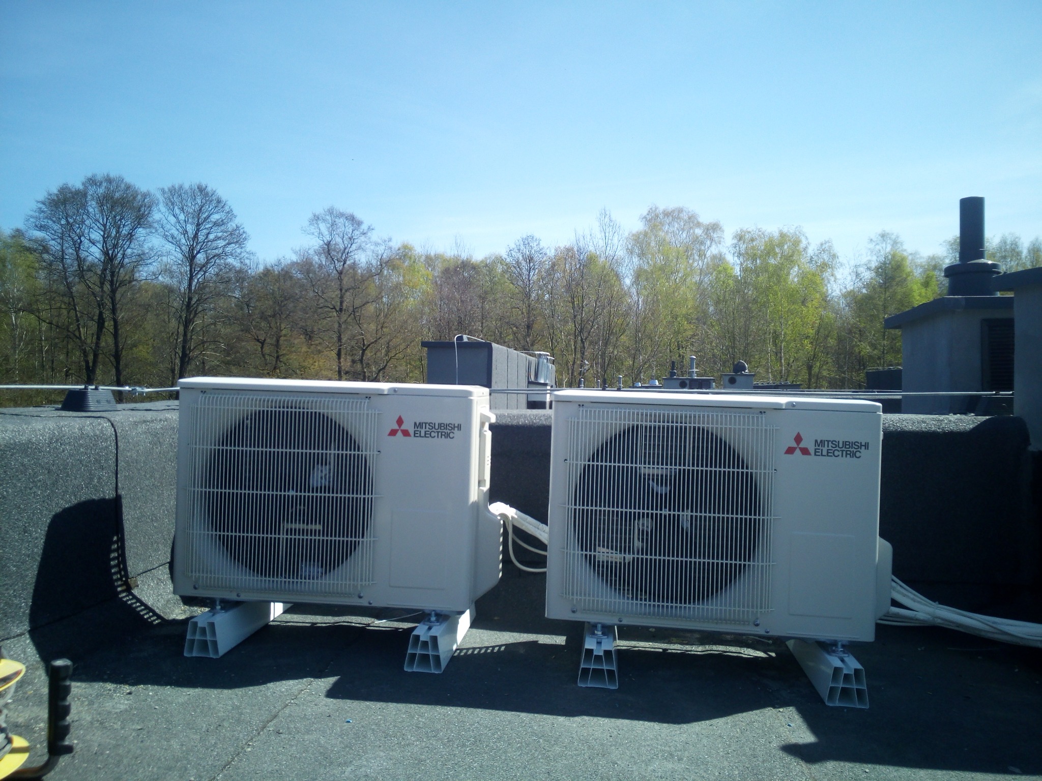 Dwa agregaty klimatyzacyjne Mitsubishi Electric zamontowane na dachu budynku, widoczne przewody i konstrukcja wsporcza, w tle drzewa i błękitne niebo.