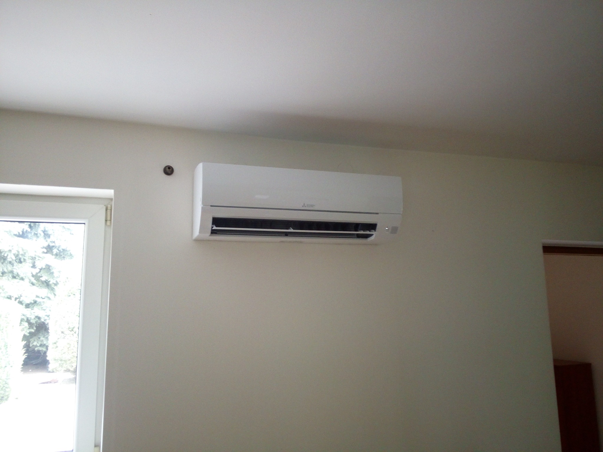 Biały klimatyzator Mitsubishi zamontowany na ścianie w jasnym pokoju, widok z boku okna.