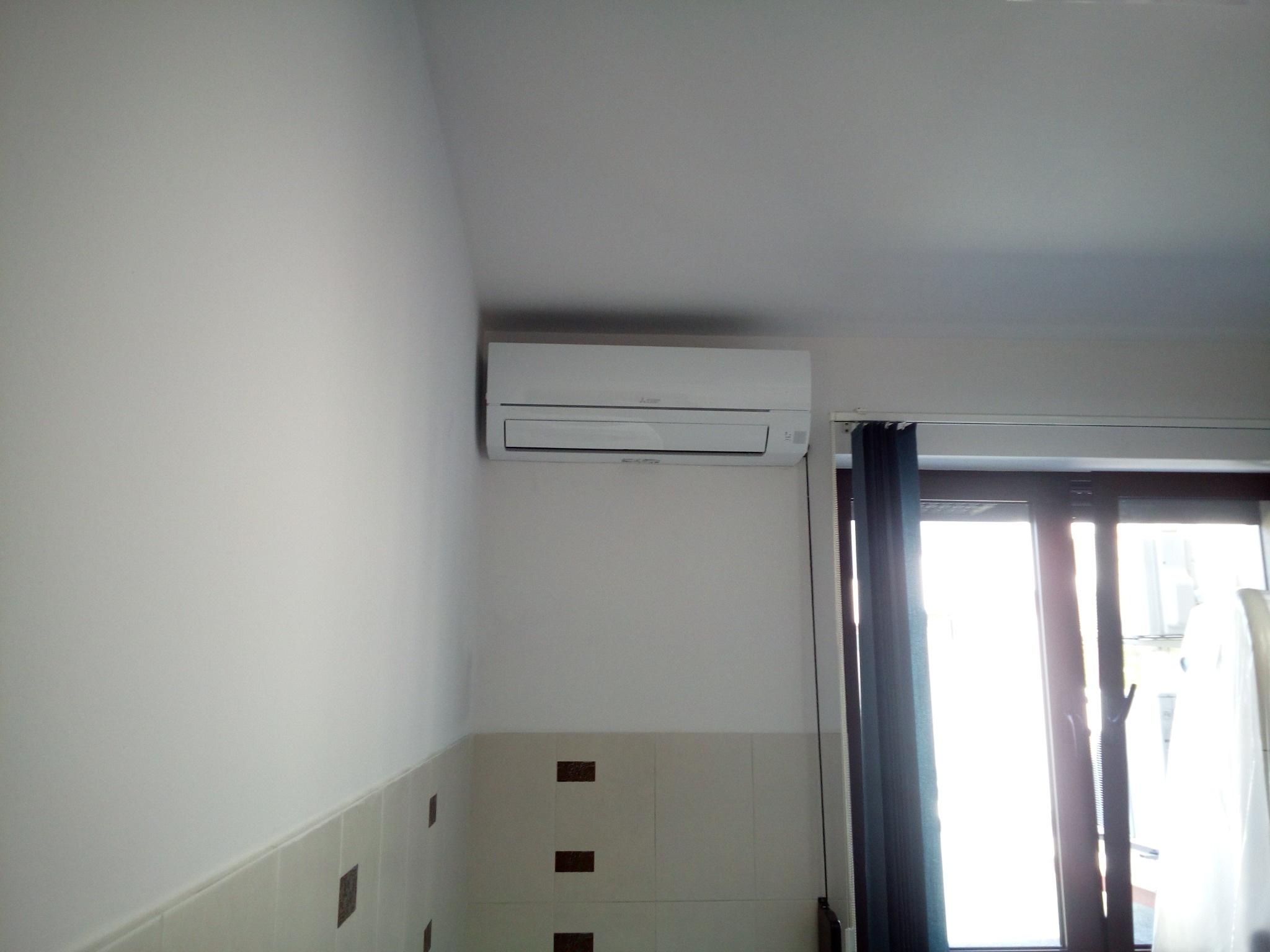 Biała jednostka klimatyzacji zamontowana na ścianie nad kafelkami w jasnym pomieszczeniu z oknem balkonowym.