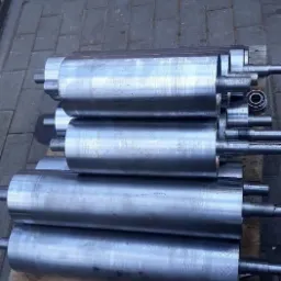 Stos obrobionych, metalowych wałków z wystającymi osiami, ułożonych na drewnianej palecie, widoczne ślady obróbki.