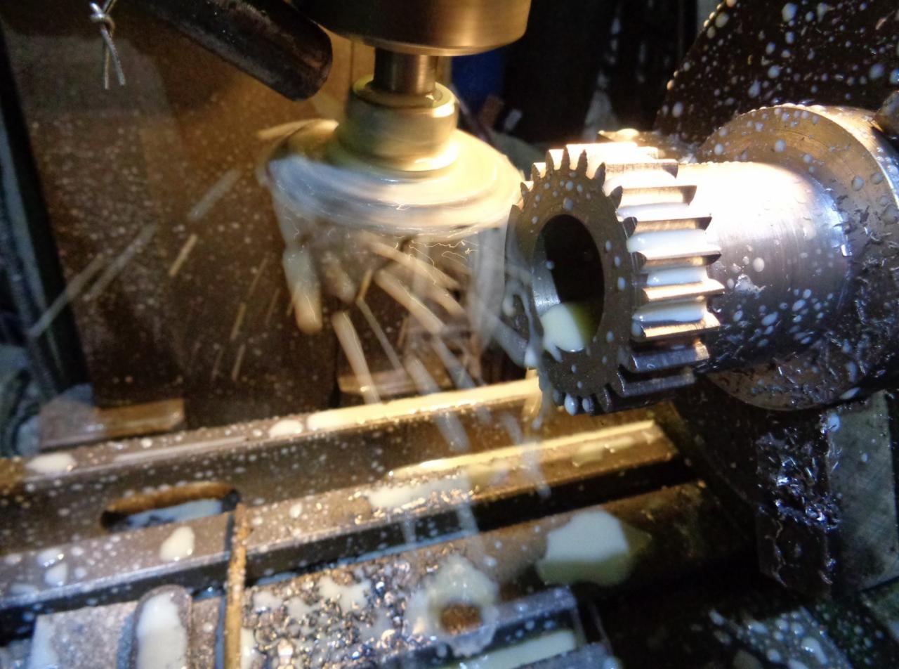 Metalowa zębatka mocowana w uchwycie jest obrabiana frezem w chłodziwie, widoczne opiłki i ciecz chłodząca.