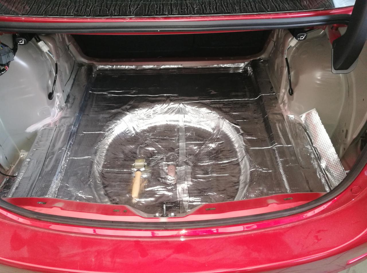 Bagażnik czerwonego samochodu wyłożony folią dźwiękochłonną, widoczny młotek z drewnianą rączką.