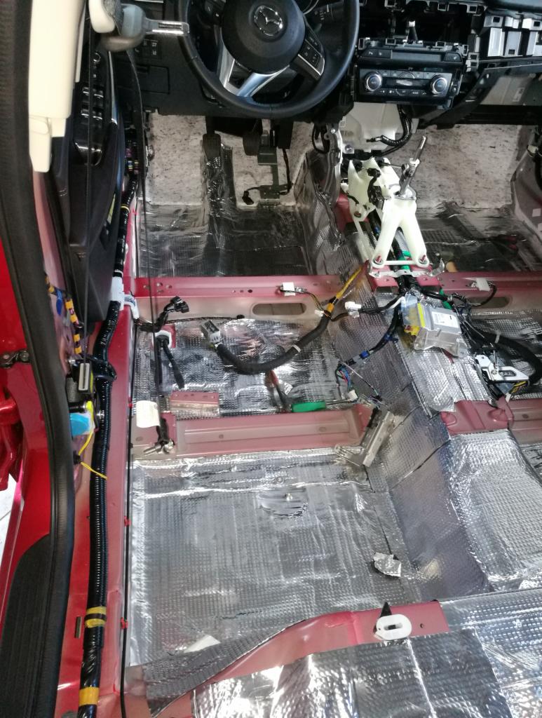 Wnętrze czerwonego samochodu w trakcie wygłuszania, widoczne maty tłumiące na podłodze i elementach karoserii, częściowo zdemontowana deska rozdzielcza.