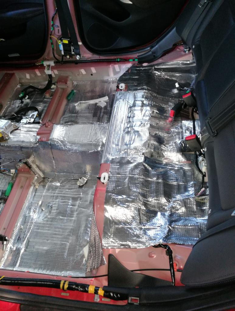 Wnętrze samochodu w trakcie wygłuszania, widoczne folie aluminiowe na podłodze, odsłonięte kable i elementy konstrukcyjne.