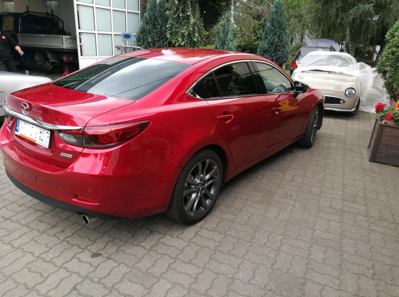 Czerwona Mazda 6 po detailingu, widoczny połysk lakieru, na tle budynku warsztatu i innego pojazdu przykrytego folią.