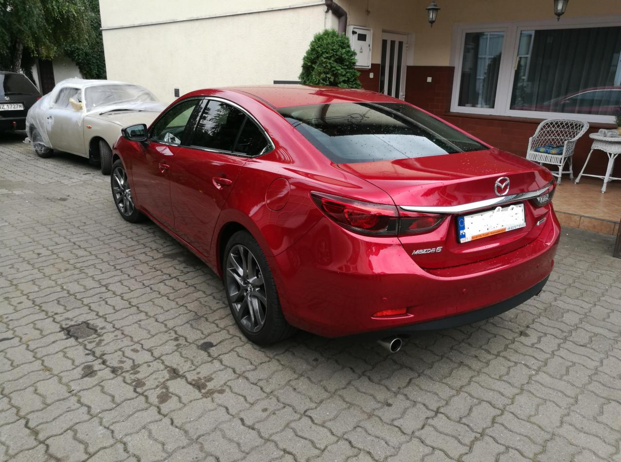 Czerwona Mazda 6 sedan z widocznymi aluminiowymi felgami, zaparkowana na brukowanej nawierzchni, w tle budynek mieszkalny i zakryty folią samochód w trakcie renowacji lakieru.
