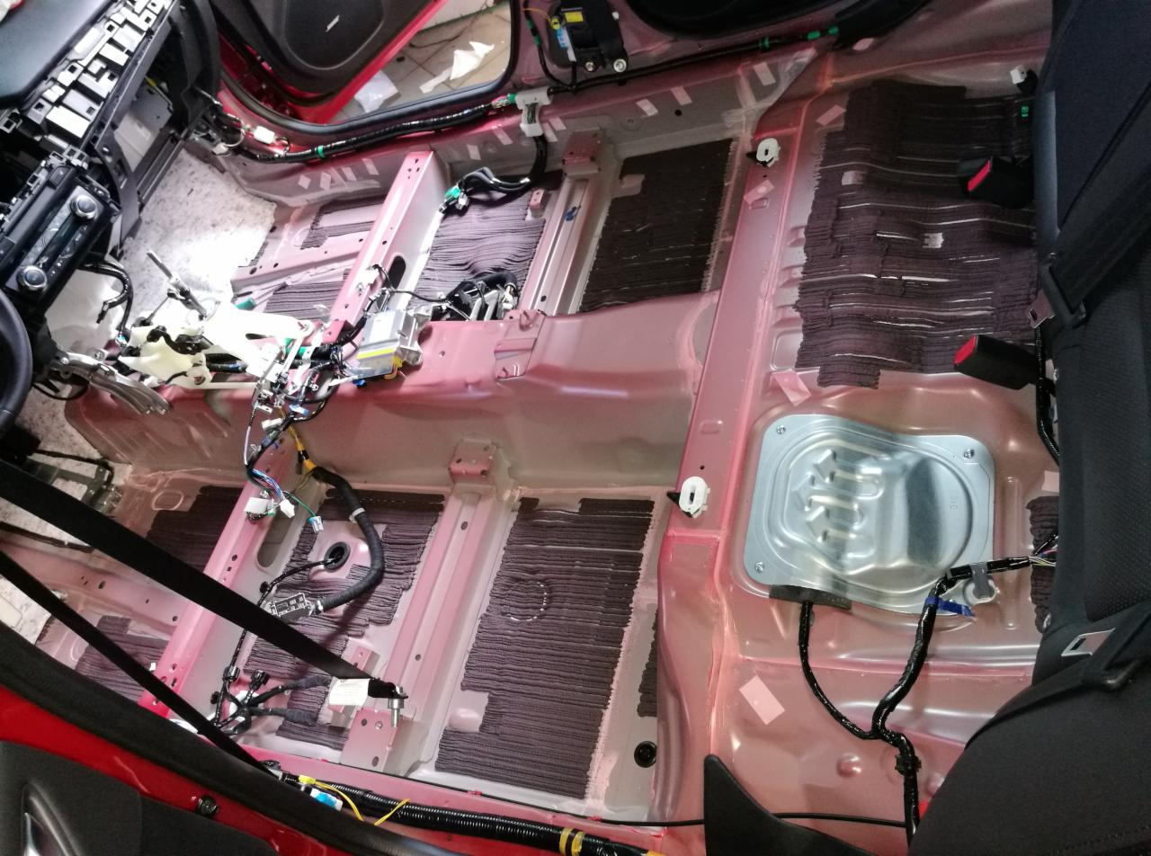 Wnętrze samochodu w trakcie demontażu, widoczne podłoga z elementami wygłuszenia, wiązki kabli, elementy deski rozdzielczej i siedzeń, czerwona karoseria.