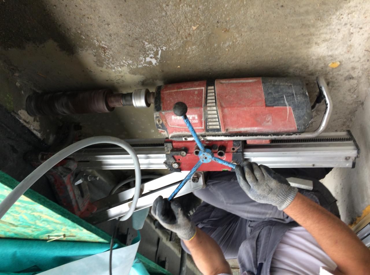 Pracownik w rękawicach obsługuje wiertnicę diamentową Hilti DD 350, wiercąc otwór w betonowej ścianie. Widoczny rdzeń wiertła i system chłodzenia wodą.