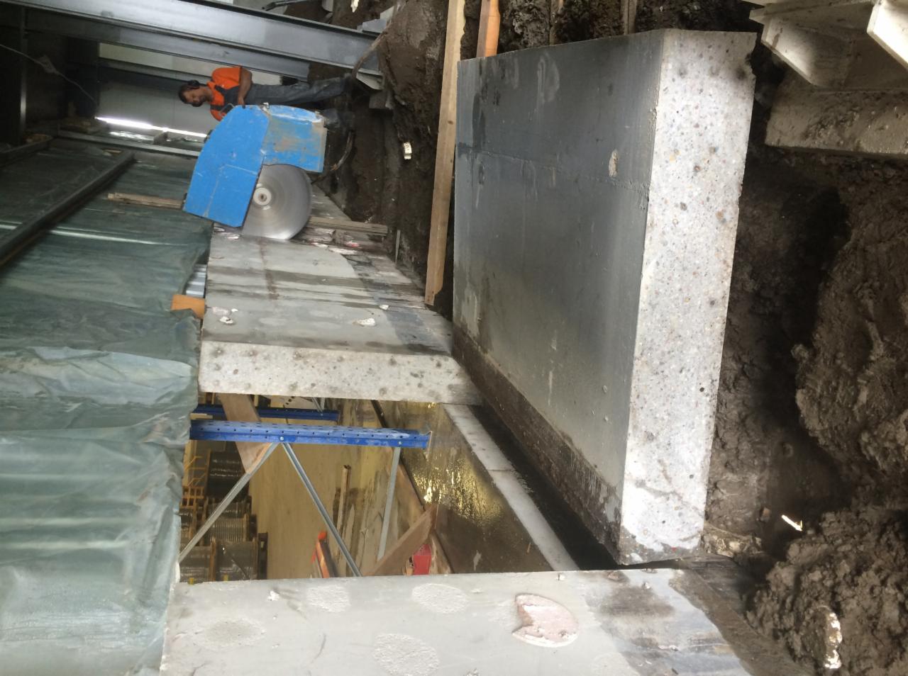 Pracownik w pomarańczowej koszulce przecina grubą betonową ścianę za pomocą piły do betonu z tarczą diamentową, widoczne fragmenty konstrukcji i ziemi.