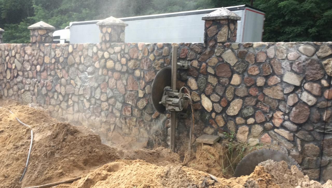 Tarcza tnąca do kamienia zamontowana na ścianie z kamienia polnego podczas cięcia, z widocznymi śladami pyłu i wykopem ziemnym u podstawy.