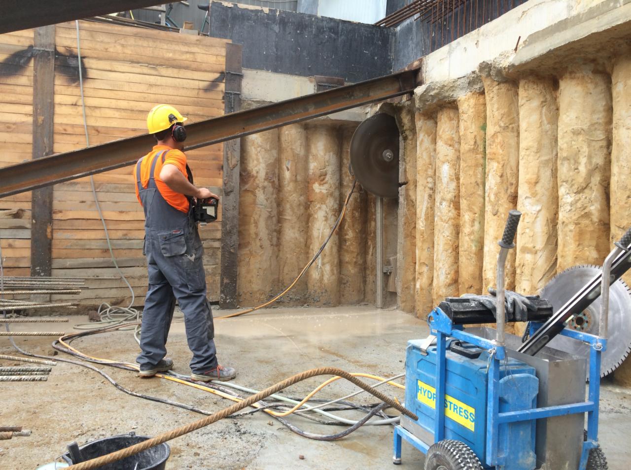 Pracownik w kombinezonie i kasku przecina ścianę z ziemi za pomocą piły diamentowej, widoczna maszyna hydrauliczna 'Hydro Stress' i stalowe pręty zbrojeniowe.
