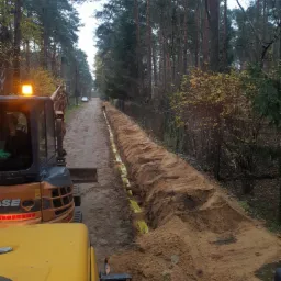 Wykopany rów z żółtą folią ochronną wzdłuż leśnej drogi, widoczny pomarańczowy mini-koparka CASE z włączonym światłem ostrzegawczym.
