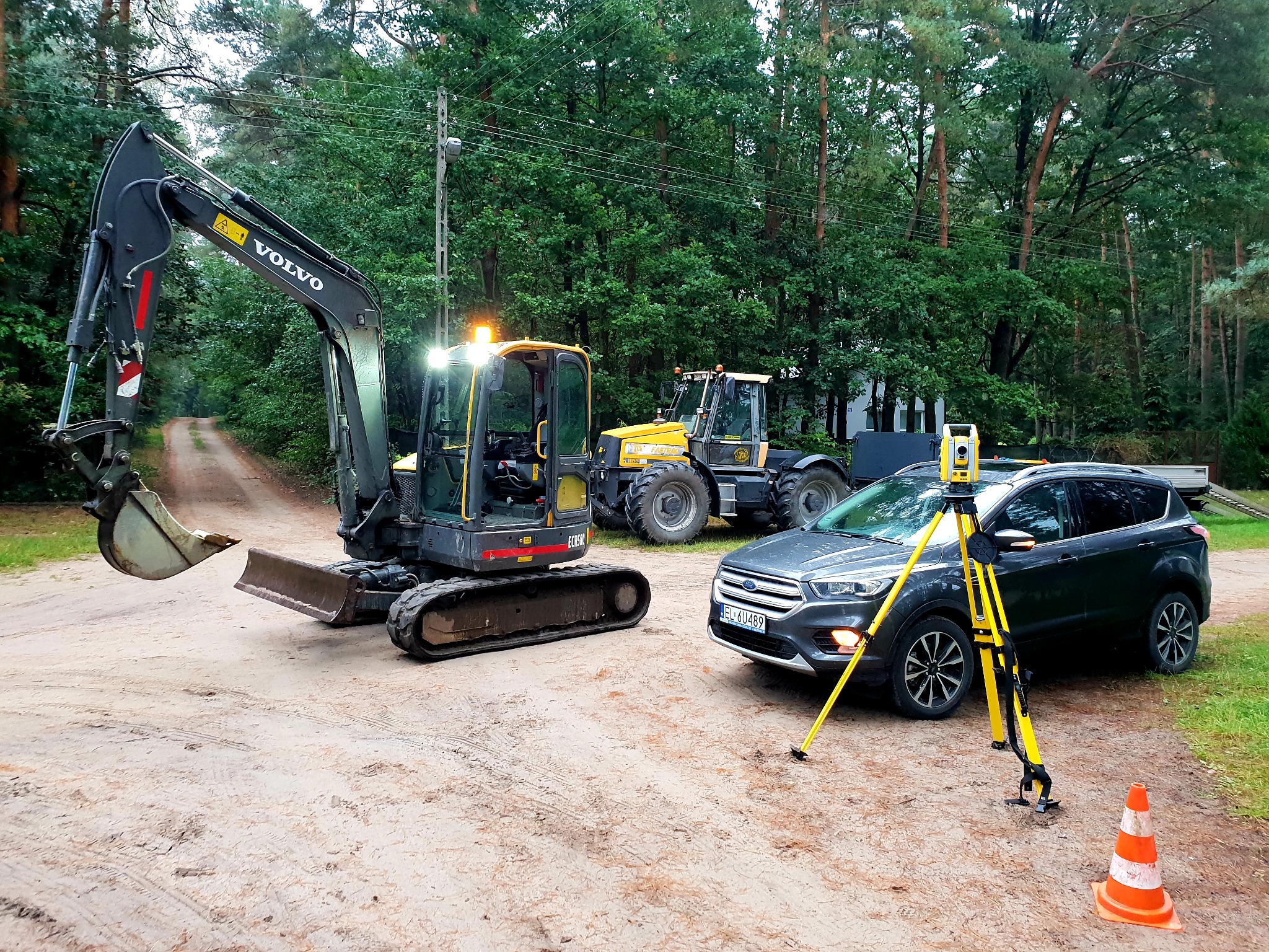 Żółty niwelator na statywie, szary samochód osobowy, czarny minikoparka Volvo i żółty traktor na piaszczystym terenie z drzewami w tle.