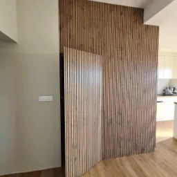 Panel ścienny z drewnianych listewek w jasnym wnętrzu, częściowo zasłaniający drzwi, widoczna podłoga imitująca drewno i fragment białej kuchni w tle.