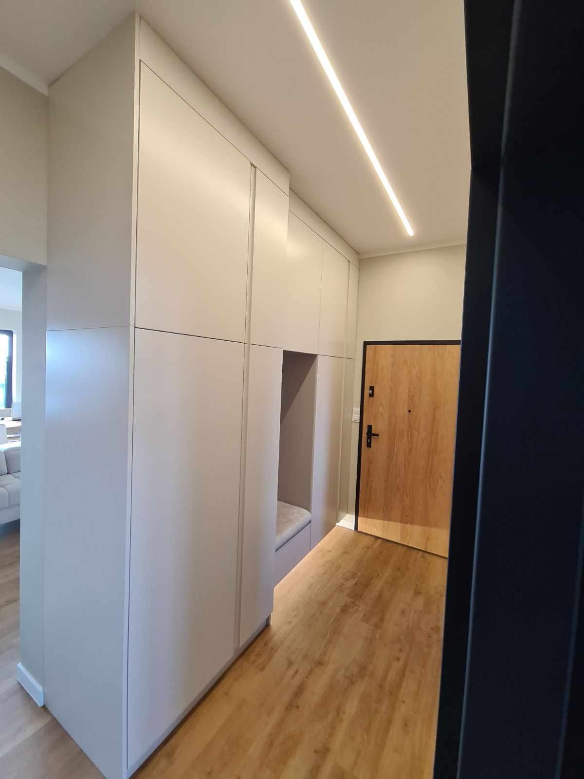 Wnętrze przedpokoju z zabudową szafy na wymiar w kolorze jasnoszarym, z wnęką do siedzenia, oświetleniem LED na suficie i drewnianą podłogą.