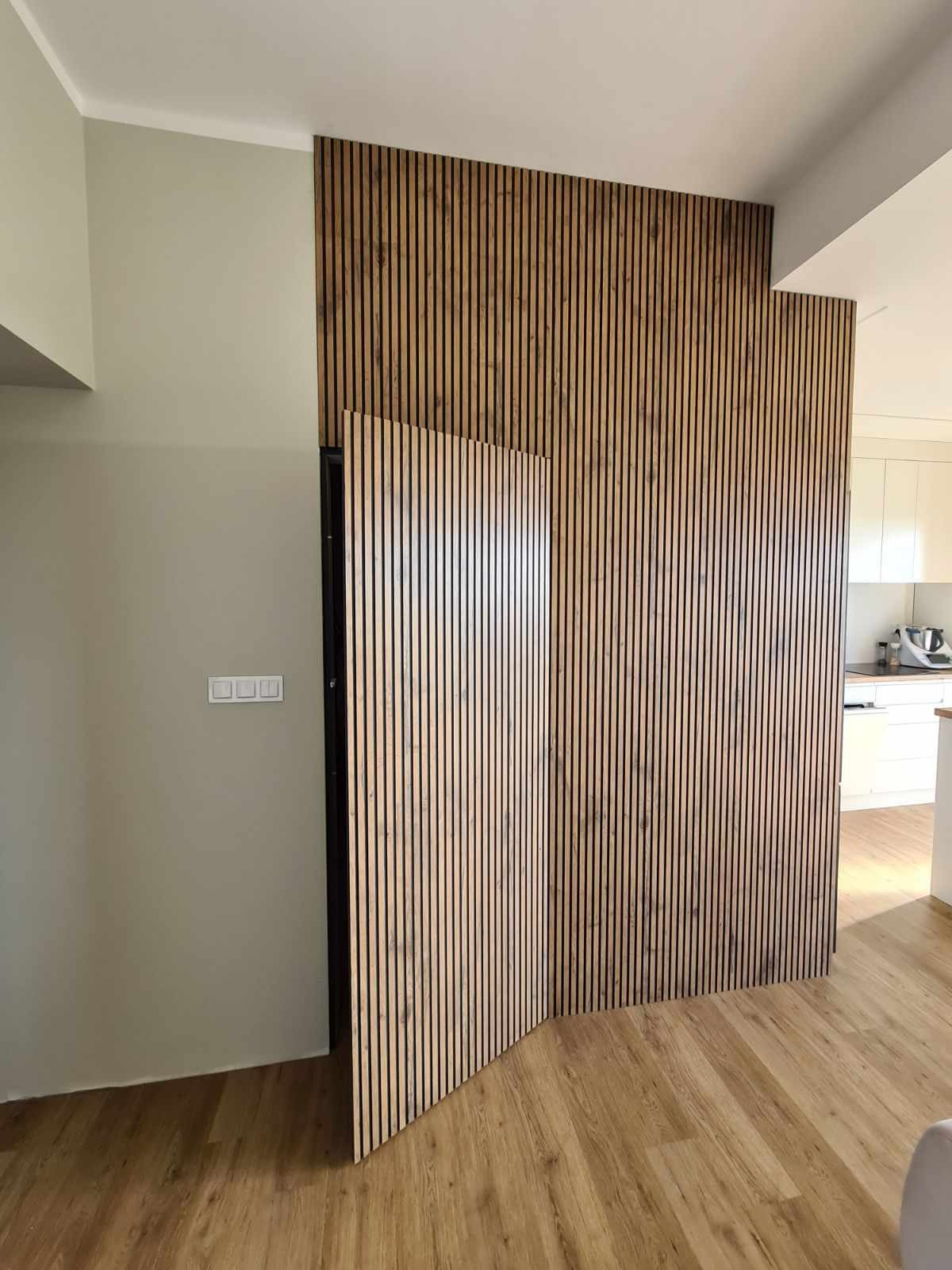 Panel ścienny z drewnianych listewek w jasnym wnętrzu, częściowo zasłaniający drzwi, widoczna podłoga imitująca drewno i fragment białej kuchni w tle.