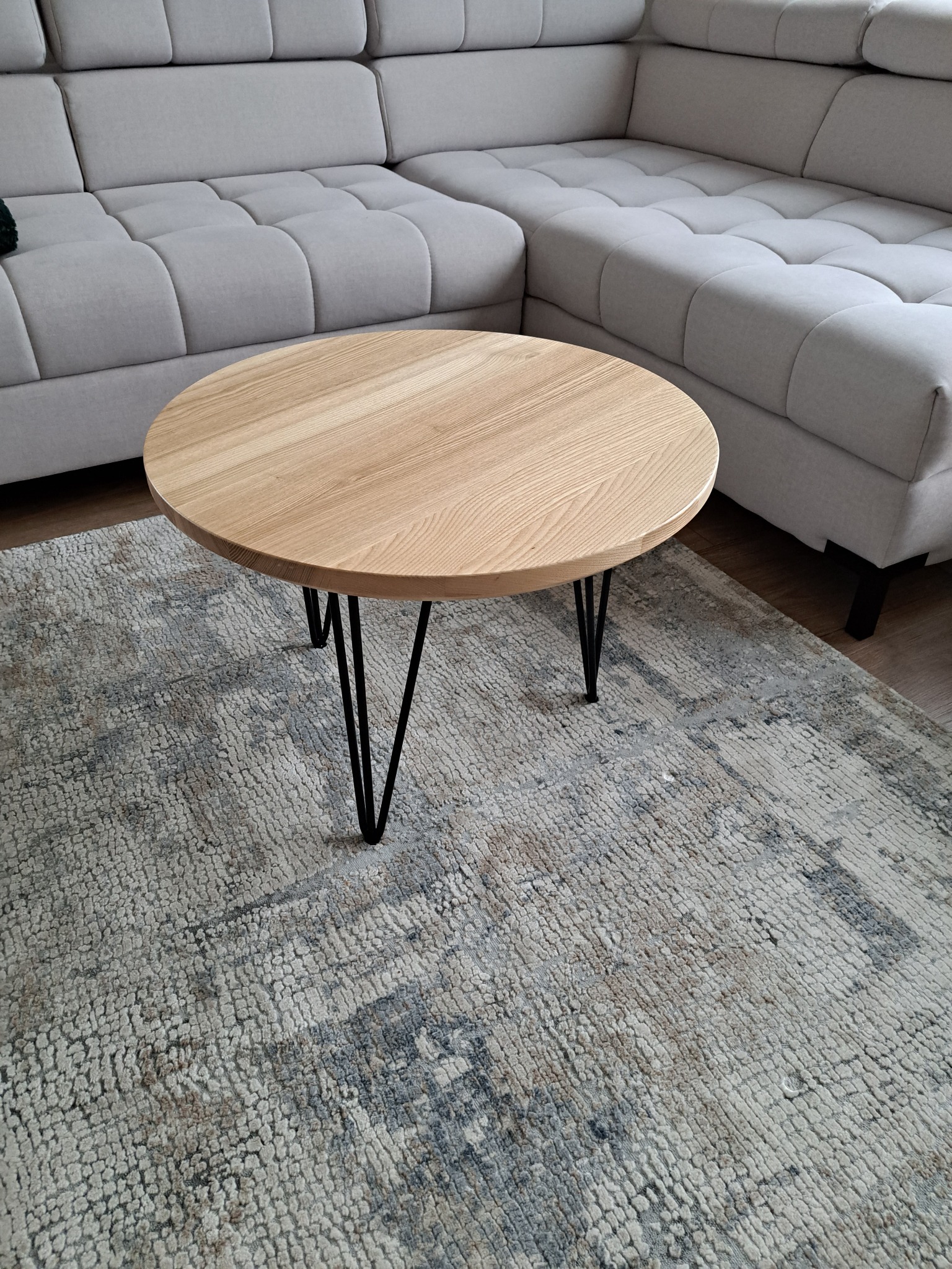 Okrągły stolik kawowy z jasnego drewna na czarnych, metalowych nóżkach w kształcie litery V, ustawiony na wzorzystym dywanie w odcieniach szarości i beżu, obok modułowej sofy z jasną tapicerką.