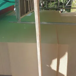 Ocieplona ściana budynku z widocznym rusztowaniem i siatką ochronną, fragment elewacji wykończony panelami imitującymi drewno.