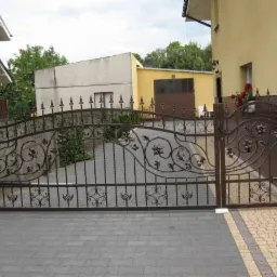 Kuta brama wjazdowa z automatyką w Kotlin, zdobiona motywami roślinnymi, prowadząca na posesję z kostki brukowej.