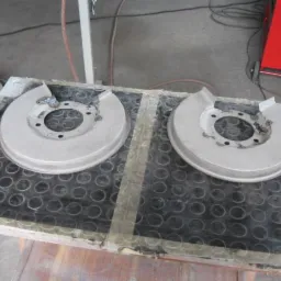 Dwa metalowe, okrągłe osłony tarcz hamulcowych, przygotowane do dalszej obróbki, leżące na czarnej, perforowanej macie w warsztacie samochodowym.