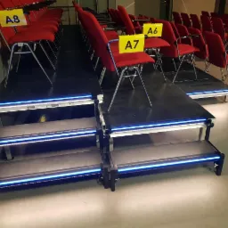 Modułowa scena z podświetlanymi schodami i rzędami czerwonych krzeseł z numeracją, widok z boku na ustawienie w sali konferencyjnej.